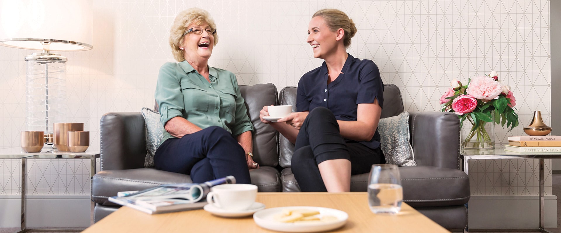 an elderly woman and her carer sitting together on a couch, laughing and drinking tea.