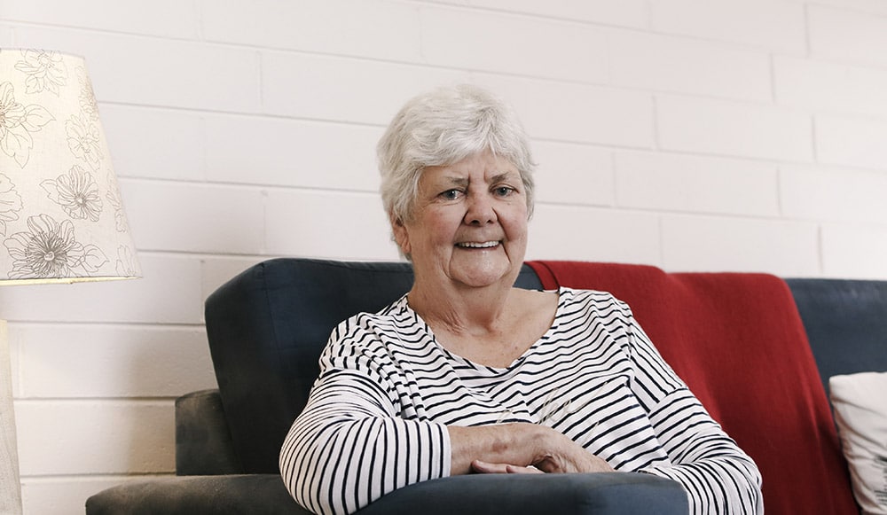 A elderly woman, sitting on a couch, smiling to camera