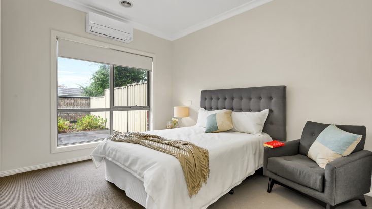 Cream coloured room with air conditioner sitting above a large window looking to the backyard, double bed with dark grey headboard is on the adjacent wall