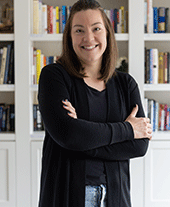 Woman with long brunette hair smiles to camera with arms crossed, white bookshelf behind her.