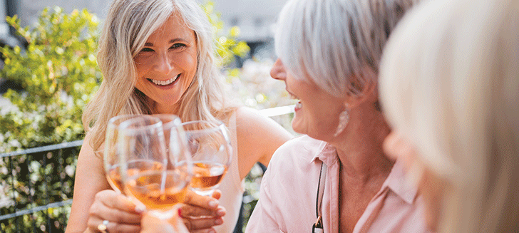 Women clinking wine glasses, both smiling, one faces the camera, the other's face is partially obscured