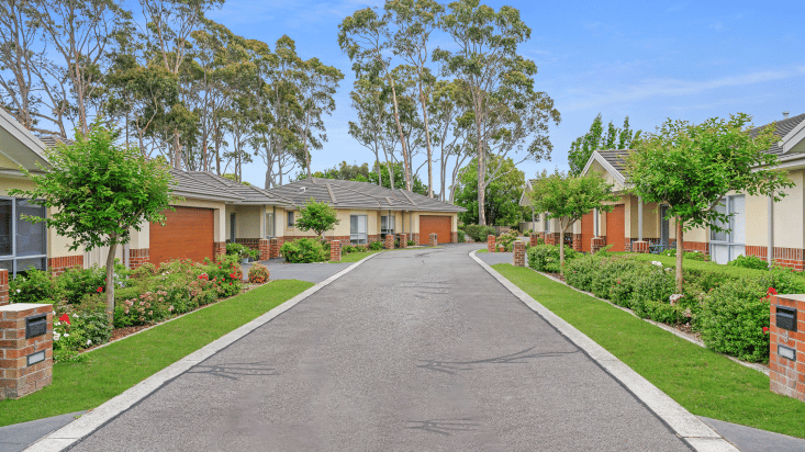 View down a tree lined street, houses are brick and cream coloured with red-brown garages