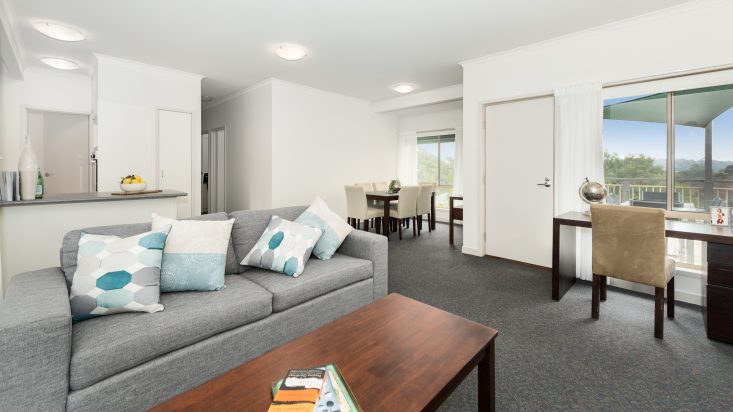 Home with white walls, grey couch and coffee table in the foreground, kitchen and dining area can be seen in the distance