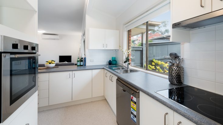 White kitchen with grey laminate countertops, dishwasher sits below the countertop and a window is above the sink