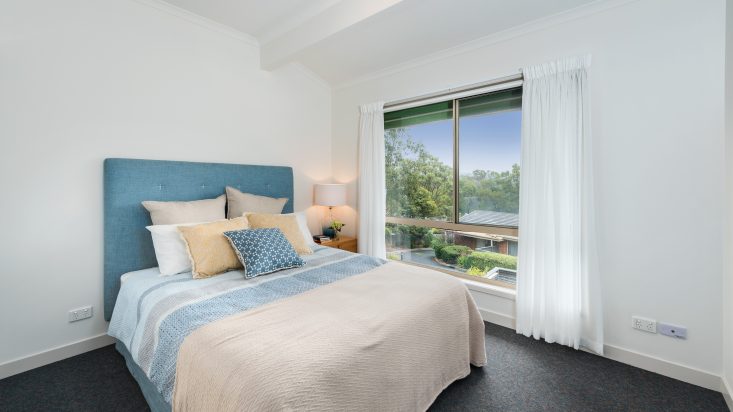 A white room with window and sheer white curtains, a bed with blue headboard sits against the adjacent wall