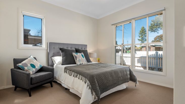 Cream coloured room with large window and ceiling fan, double bed with grey bedhead sits against adjacent wall, grey armchair sits next to the bed