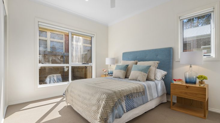 Cream coloured room with ceiling fan, a large window looks out to the fence in the backyard, a double bed with blue headboard sits on adjacent wall