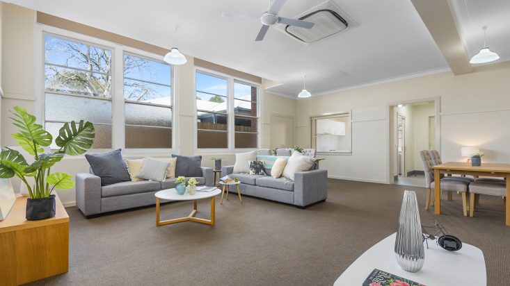 Large room with ceiling fan and brown carpet, two grey couches sit in the centre of the room around a round coffee table, large windows adorn the wall behind