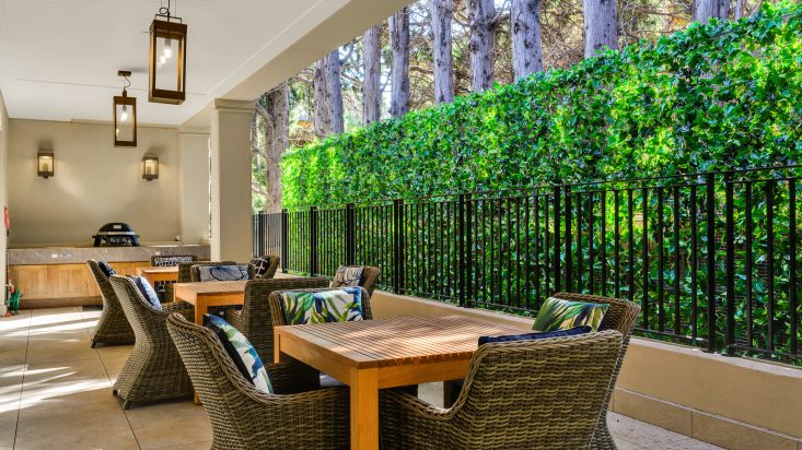 Wicker dining sets sit outside underneath a covered patio, green hedge offers privacy to the right