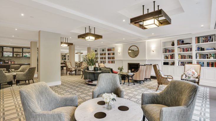 Grey chairs sit around a white marble tables, armchairs and lounges can be seen against the far wall containing a bookshelf