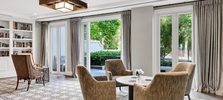 Grey armchairs sit around a marble topped table, a wall of full length windows sits behind, one is open showing the greenery outside