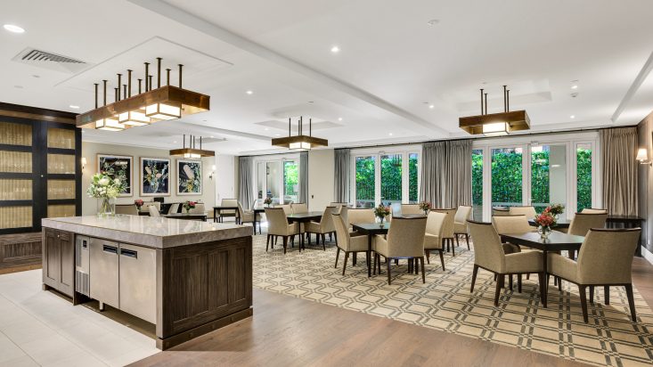 Walnut dining tables with cream coloured chairs fill a white room with patterned cream carpet floors, a kitchen can be seen to the left