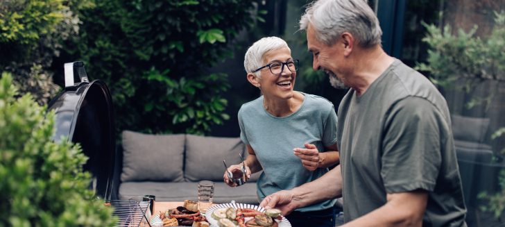 Older woman and man outside barbequing, food is on the grill and greenery is behind them.