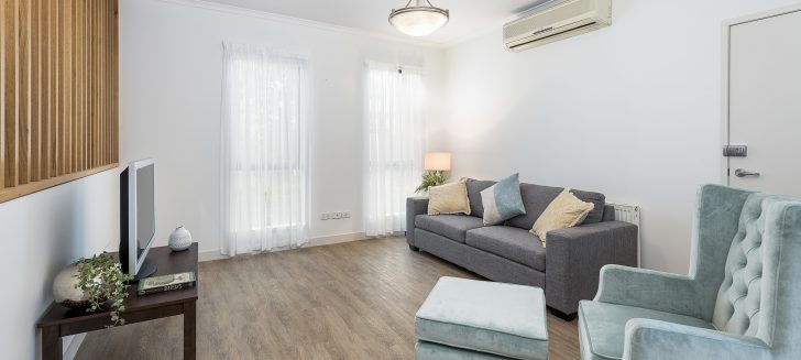 White lounge room with curtains covering the windows. A grey couch and light blue arm chair sit on a wooden floor with the tv opposite.