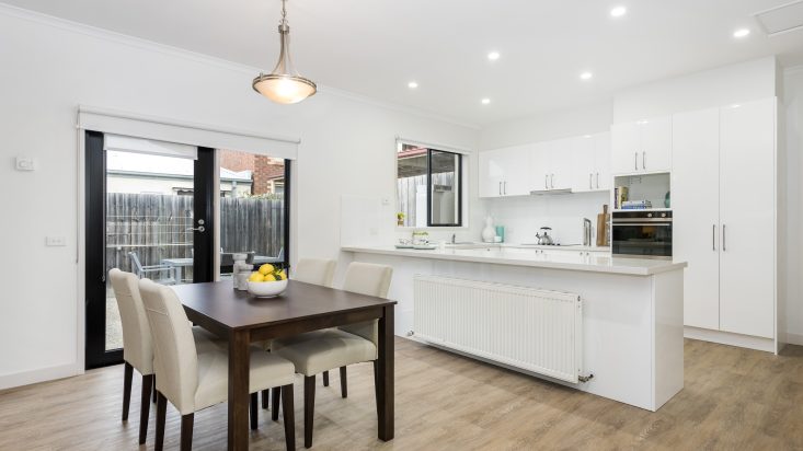 Wooden dining table with white chairs sits on a wooden floor next to sliding doors, white kitchen in the background.