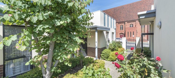 Walkway with shrubs and trees in front of a cream and brick building, pedestrian gate to right hand side