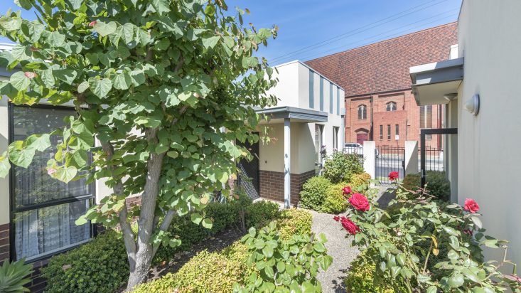 Walkway with shrubs and trees in front of a cream and brick building, pedestrian gate to right hand side