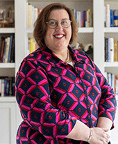 Woman with short brown hair and glasses wears a navy blouse with bright pink pattern smiles to camera, white bookshelf behind her.