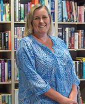 Woman with shoulder length blonde hair in blue patterned blouse smiles to camera, light wooden bookshelf behind her.