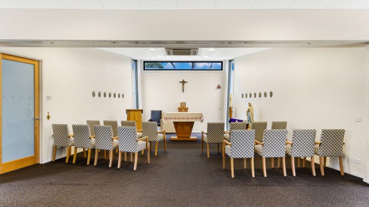 Two rows of patterned chairs sit in front of a Catholic altar, cross on back wall