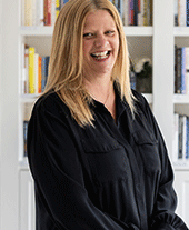 Woman with long blonde hair and black button up shirt smiles to camera, white bookshelf behind her.