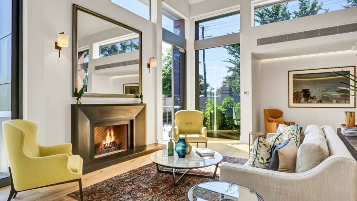 Grey lounge and yellow armchairs around a metal fireplace, tall windows on adjacent walls