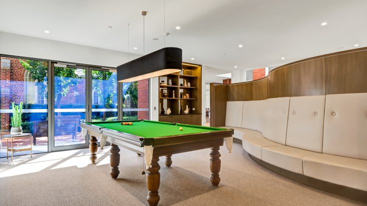 Pool table with large black pendant light above, full length sliding doors lead outside and sit next to a walnut bookshelf in the background