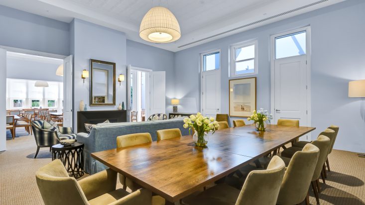 Room with light blue walls has a large wooden dining table in foreground, a blue lounge can be seen behind