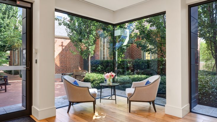 Grey armchairs sit in front a corner window facing each other, pink flowers sit on the side table in the centre