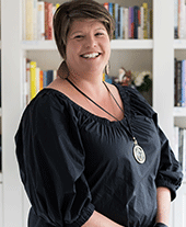 Woman with short brown hair and black blouse smiles to camera, white bookshelf behind her.