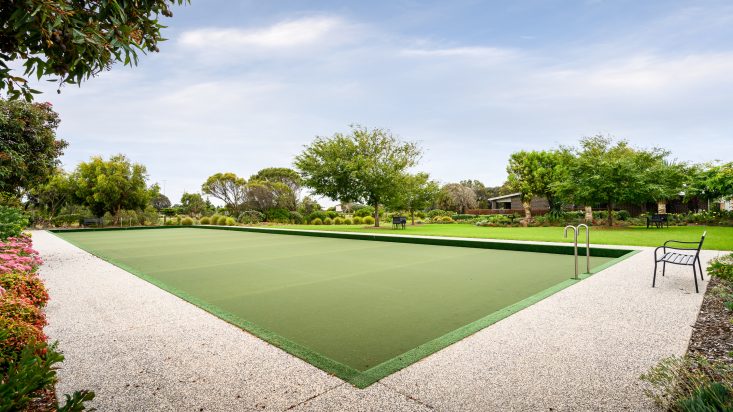 Outdoor bowling green on a sunny day, trees and shrubs behind it.