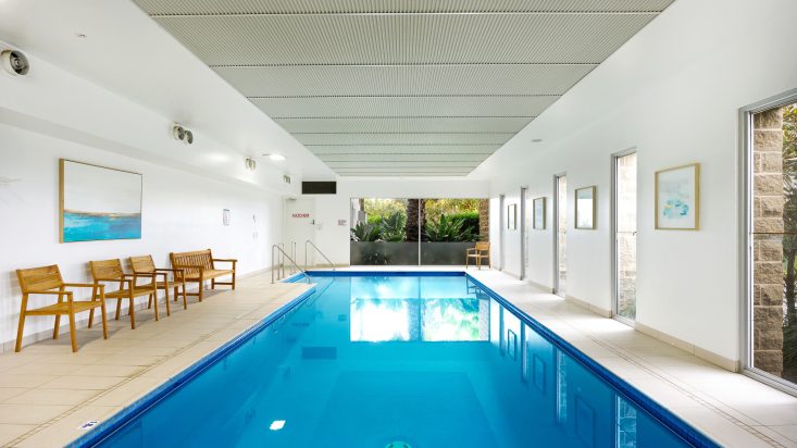 Deep blue indoor pool in white room with a large window looking out to greenery. Chairs can be seen to the left.