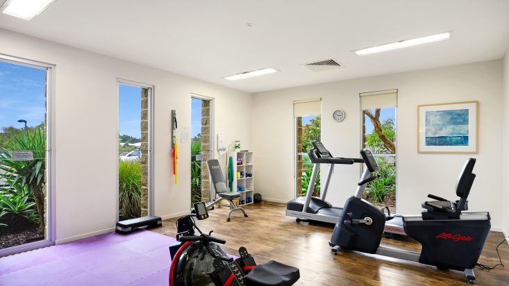 Gym with white walls and wooden floor, greenery is visible outside the 4 tall windows. Yoga mat, treadmill, fitness bench, and elliptical are in view.