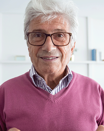 Man half smiles to camera, wearing glasses and a purple knit