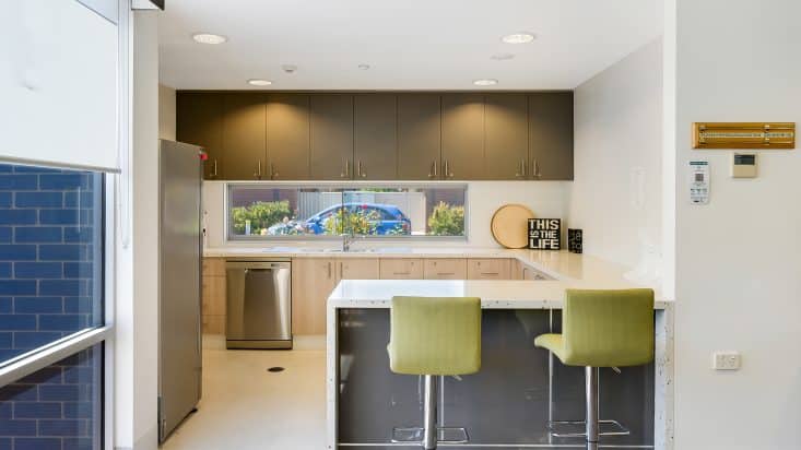 Green bar stools sit under a U-shaped kitchen bench, stainless steel fridge fridge and dishwasher can be seen in background