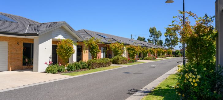 A row of white and brick houses to the left of the street with short trees in front yards, lamp post on the opposite side