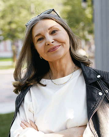 Woman smiles to camera with mouth closed, greenery in background