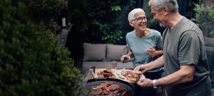 Older woman and man outside barbequing, food is on the grill and greenery is behind them.