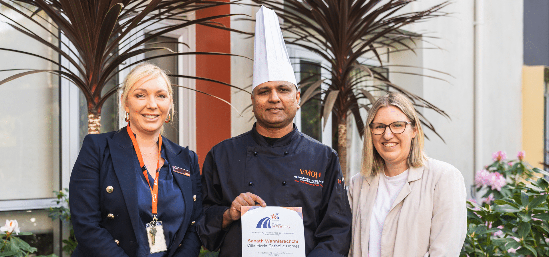 A chef stand with two women. He is holding a certificate.