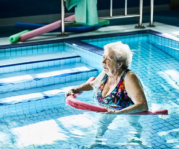 An elderly woman in a pool, walking with a pool noodle