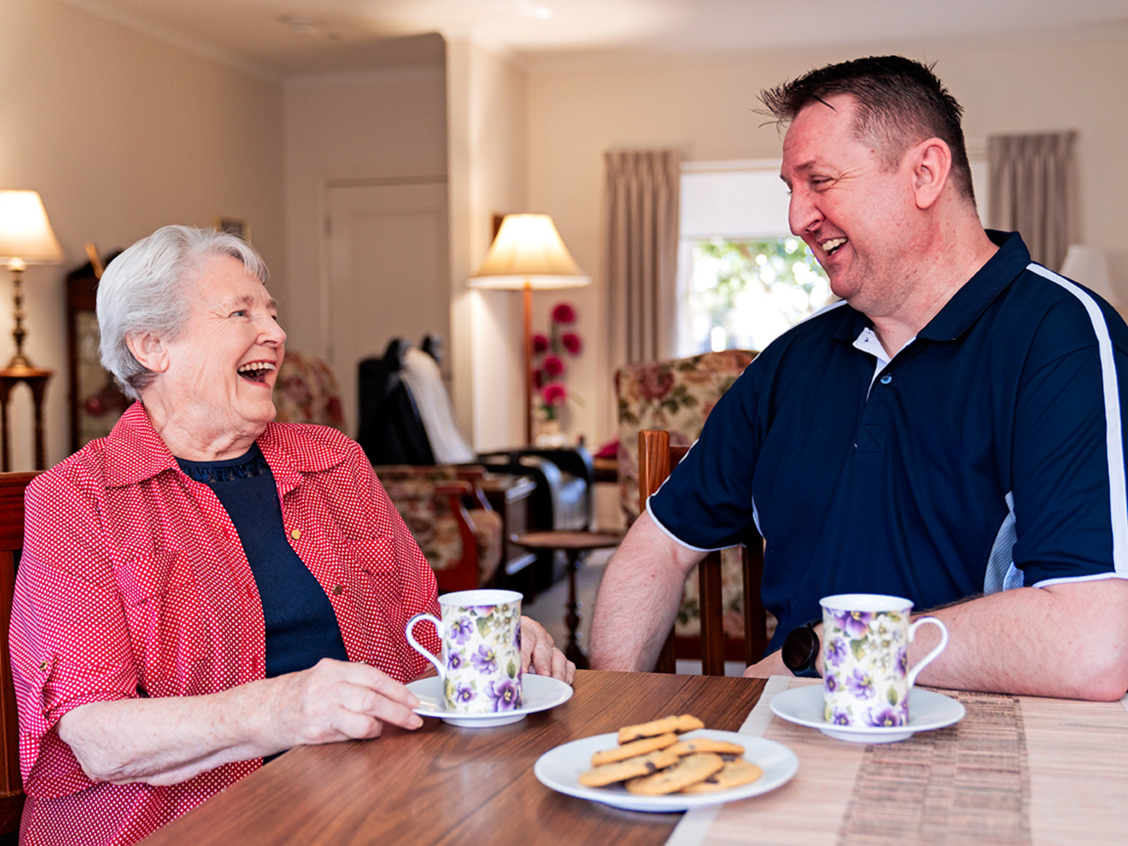 An older woman and a man share a laugh. They have a cup of tea and biscuits in front of them on a table.