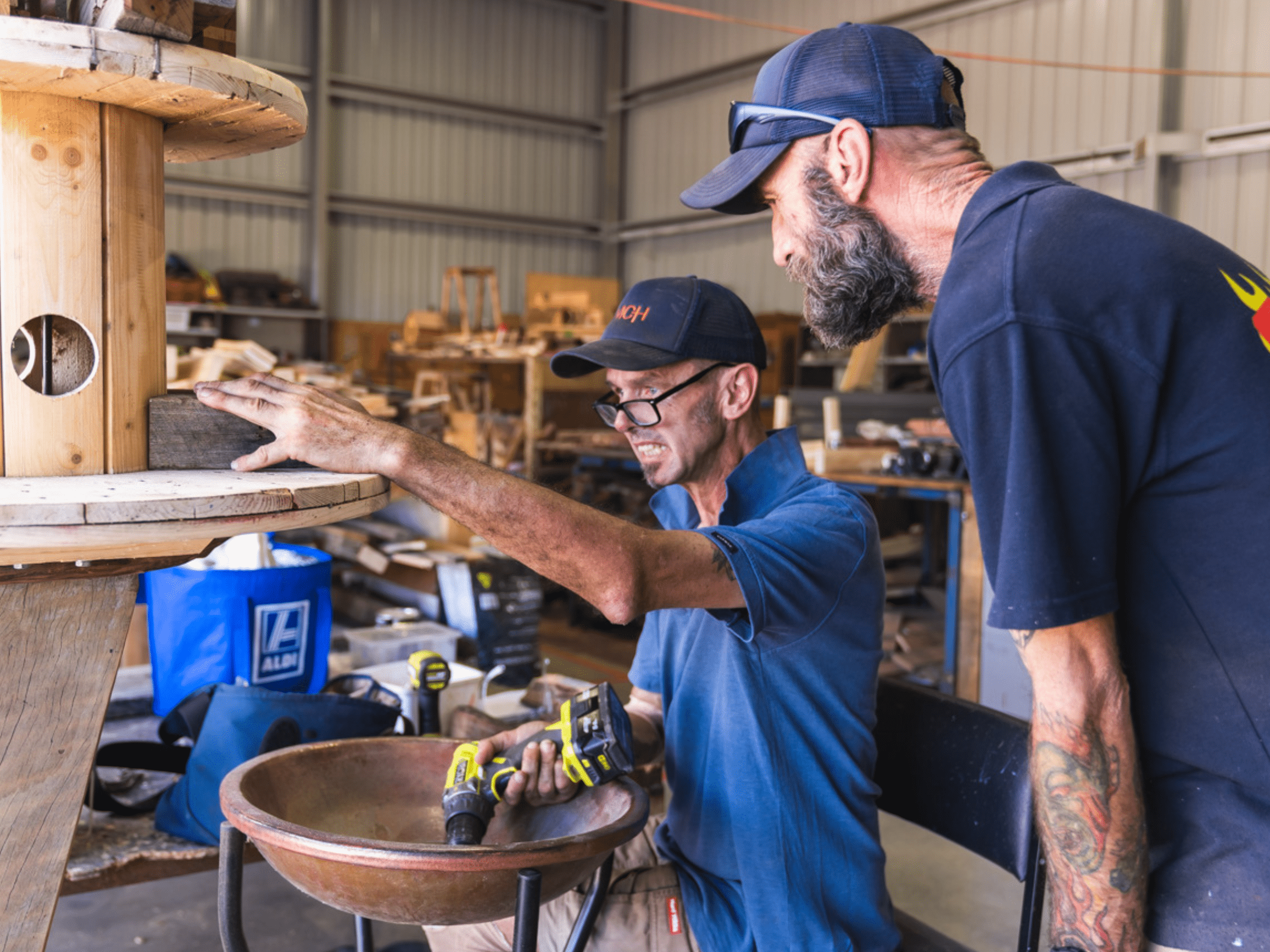 A man uses tools to work on a bird bath. A second man stands next to him.