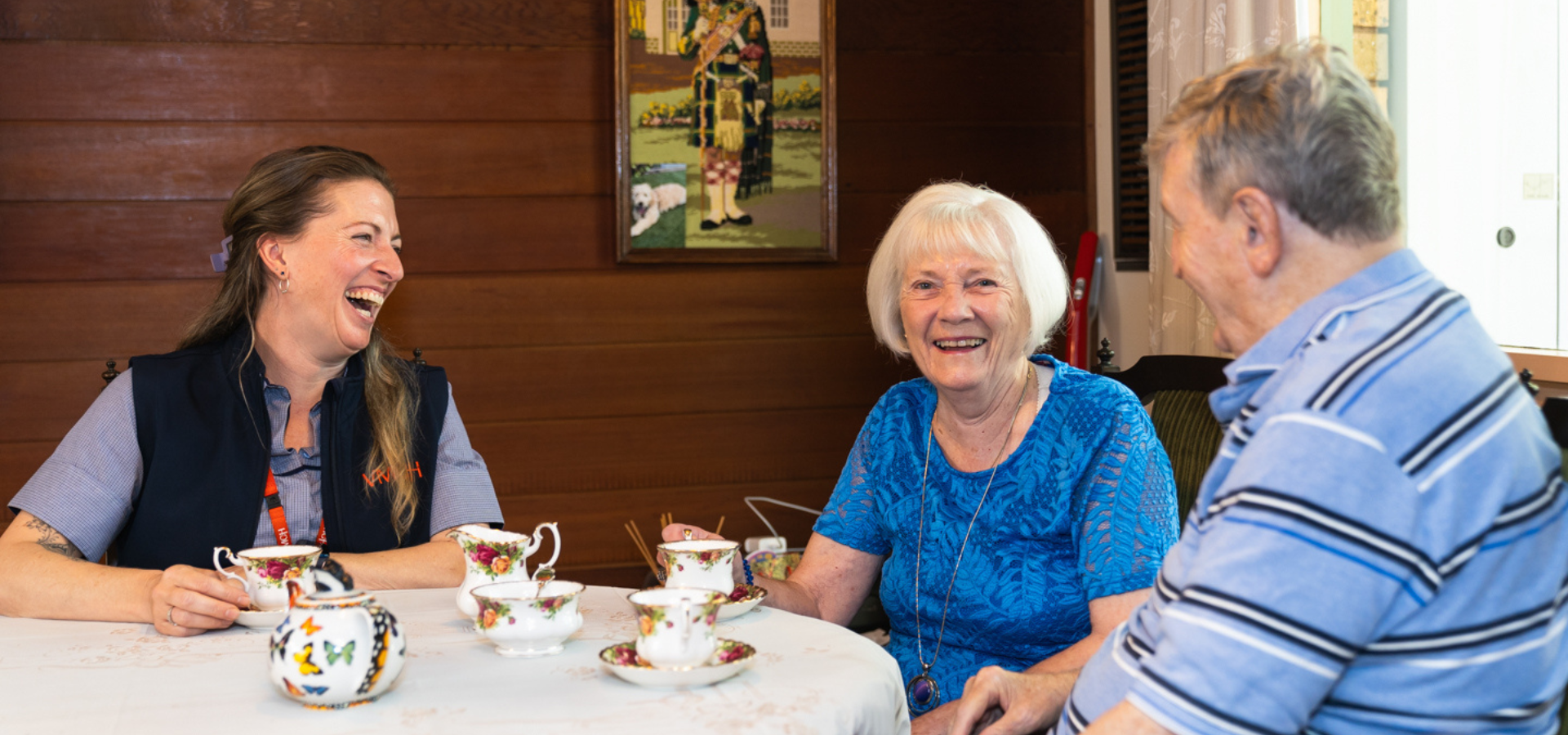 An older man and woman sit at a table with a younger woman. They are smiling and having a cup of tea.