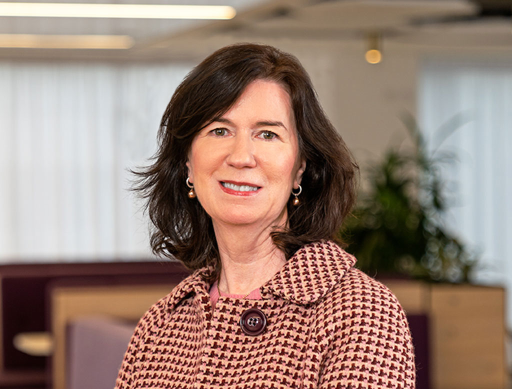 A woman wearing a coat, standing in a corporate office, smiling.