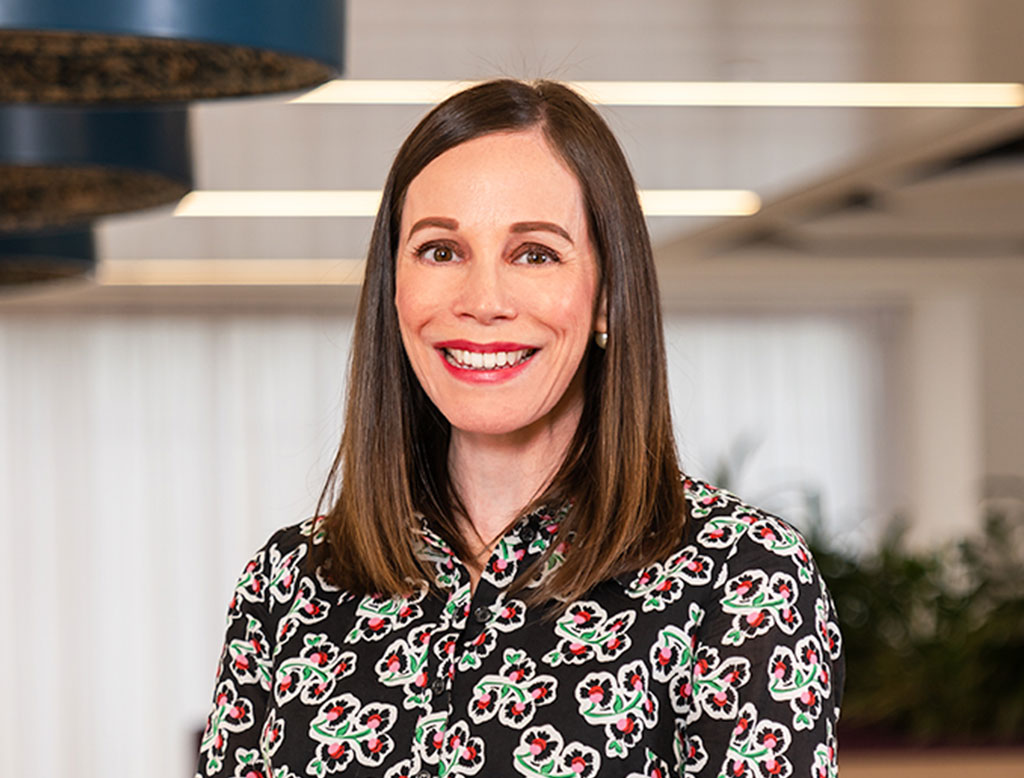 A woman wearing a black floral shirt, standing in a corporate office, smiling.