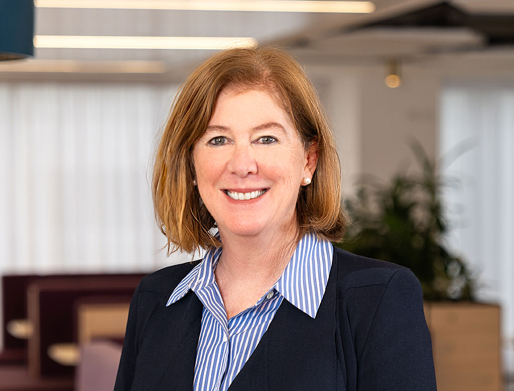 A woman wearing a blue shirt and jacket, standing in a corporate office, smiling.