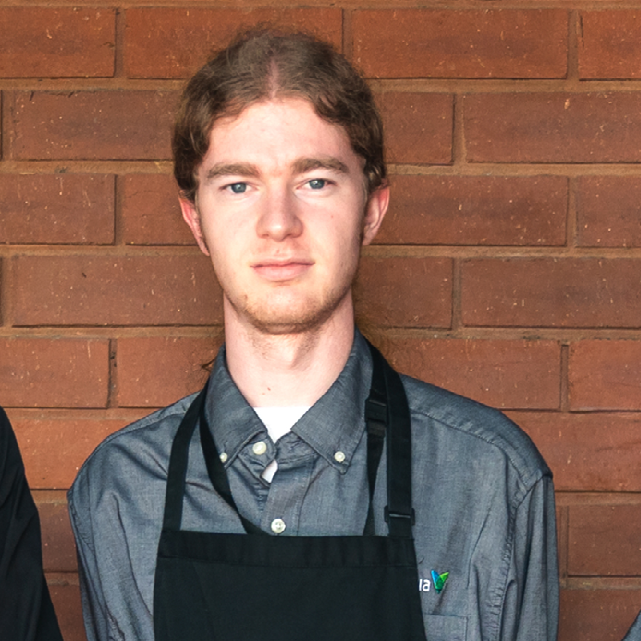 Man standing in front of brick wall wearing a grey shirt and black apron