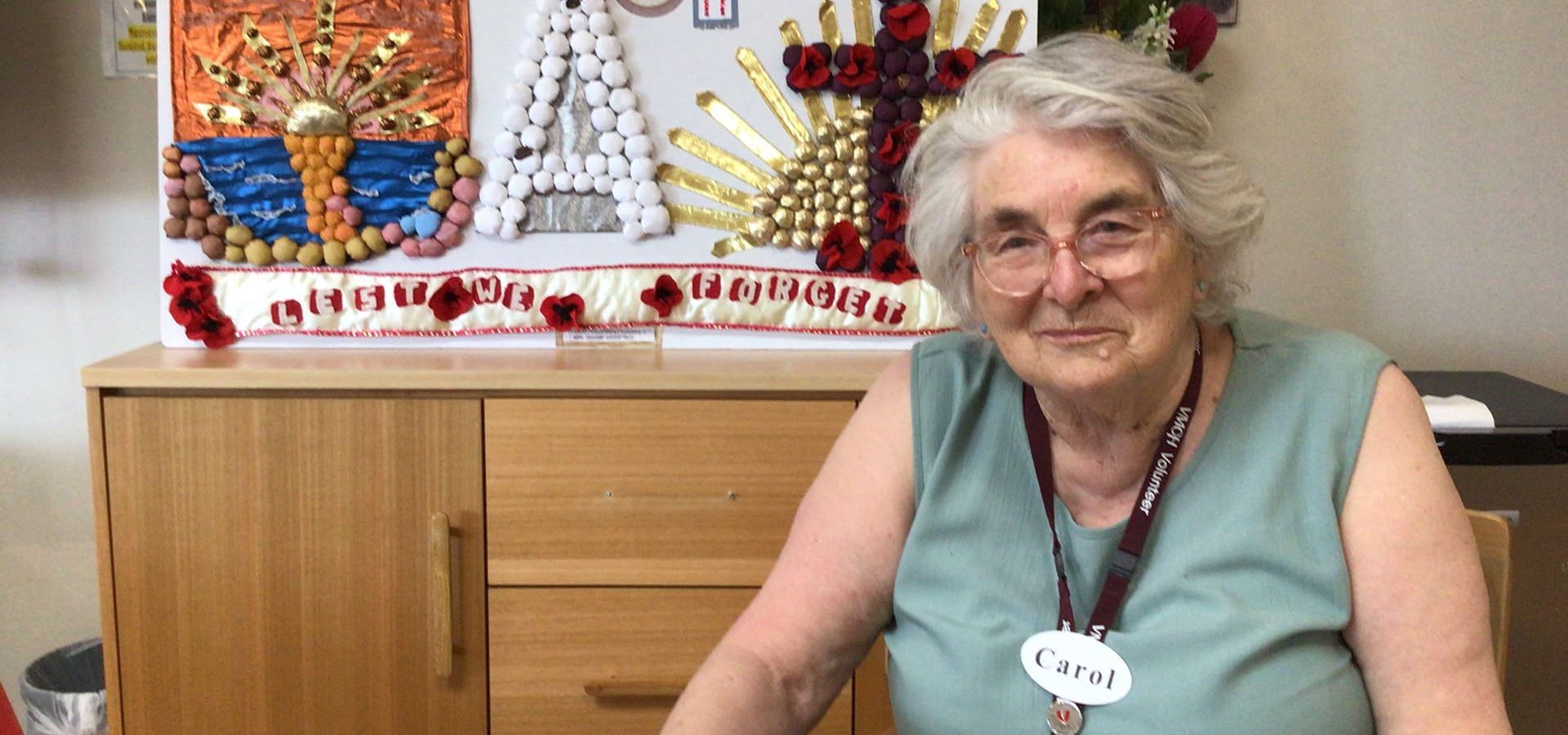 A woman wearing a green shirt and a nametag that reads 'Carol' sits at a table and smiles at the camera