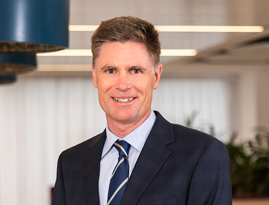 A man wearing a suit, standing in a corporate office, smiling.