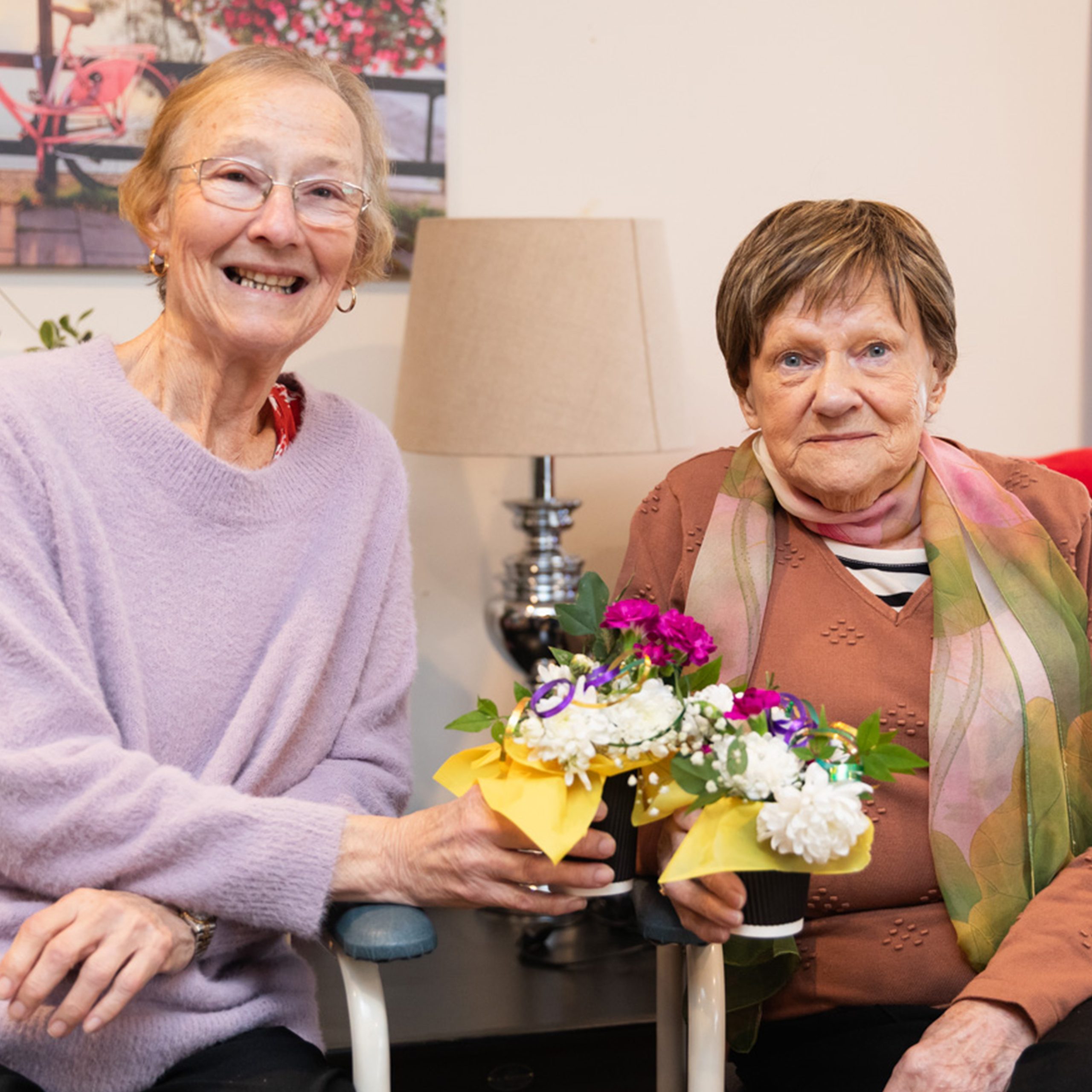 Two woman in their seventies smiling and showing bouquets of flowers they made.